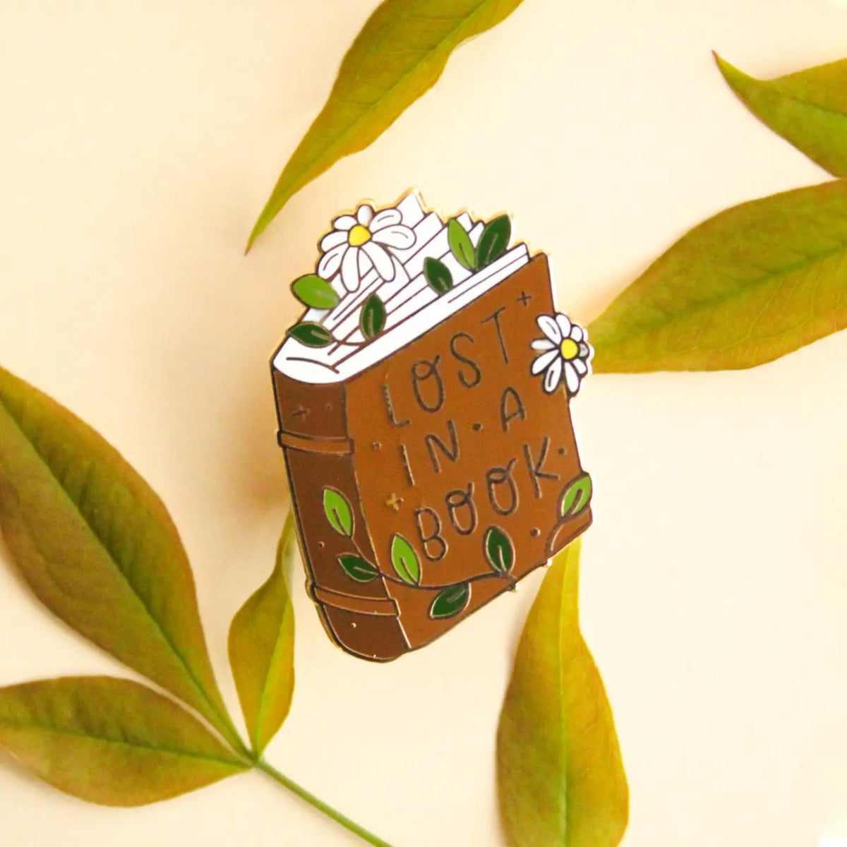 Pin's émaillé Occasionalish en forme de livre fleuri de marguerites avec le message Lost in a Book
