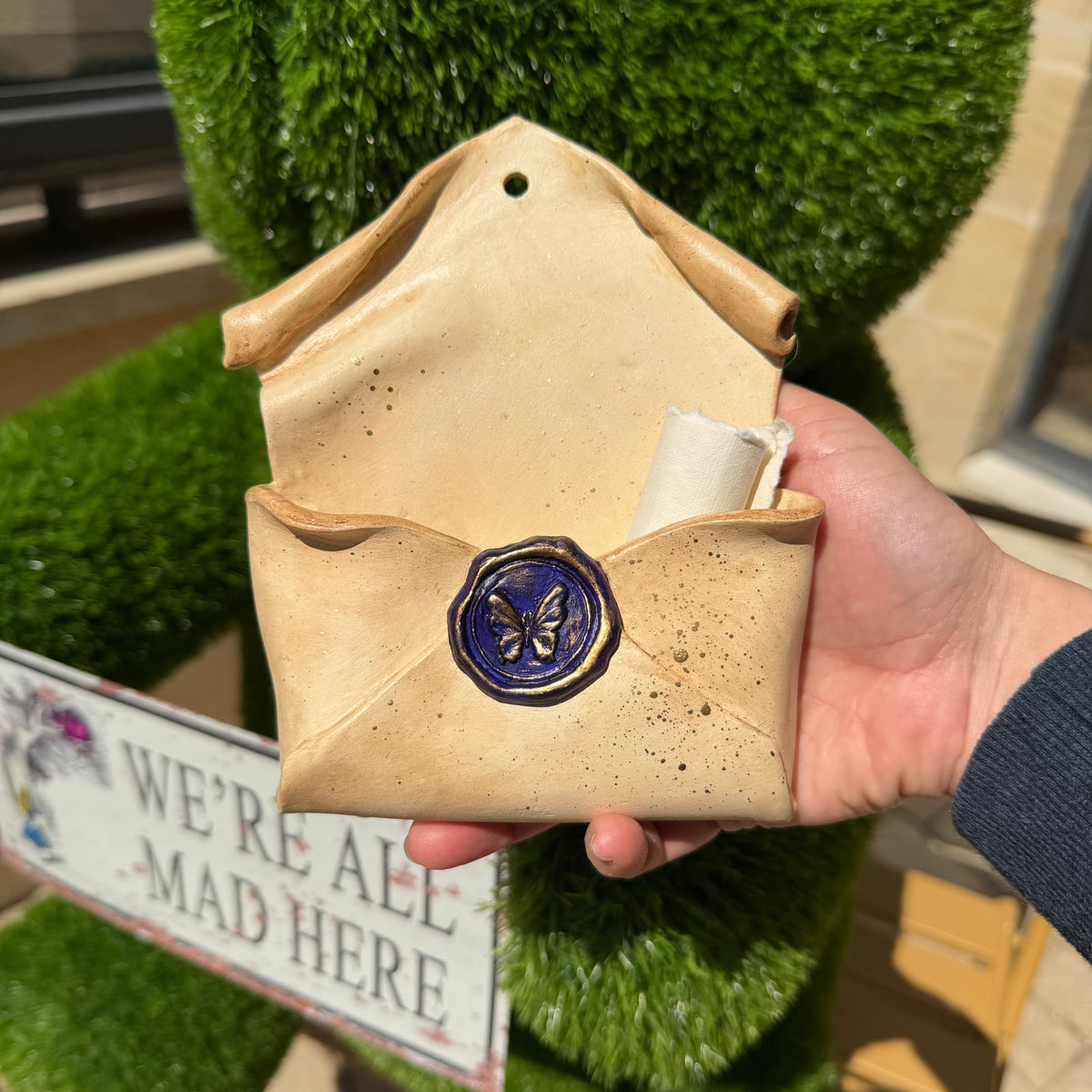 Enveloppe murale artisanale en argile effet parchemin fermée par un sceau violet et doré orné d'un papillon