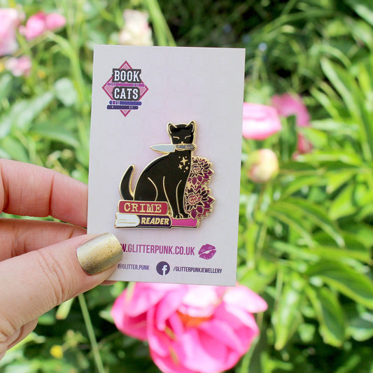 Pin's Glitter Punk Crime Reader, chat noir avec couteau assis sur une pile de polars et fleurs pourpres