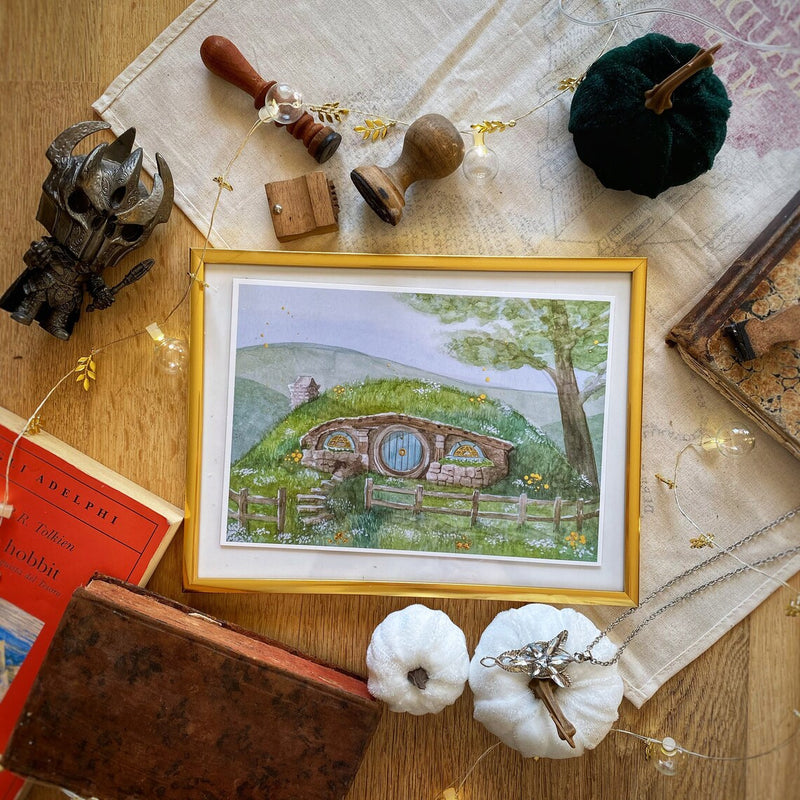 Illustration A5 d'une maison de hobbit à porte ronde et toit végétalisé nichée dans les collines verdoyantes